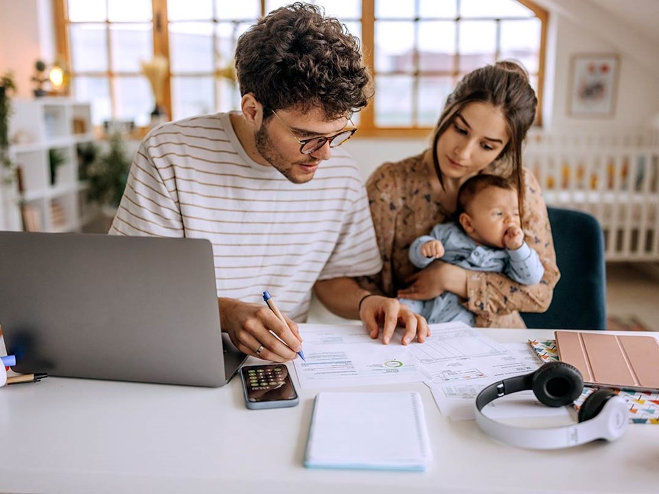 Mann jobber seg gjennom dokumenter som ligger på et bord med en kvinne og en baby vedsiden av, til illustrasjon for økonomiske problemer
