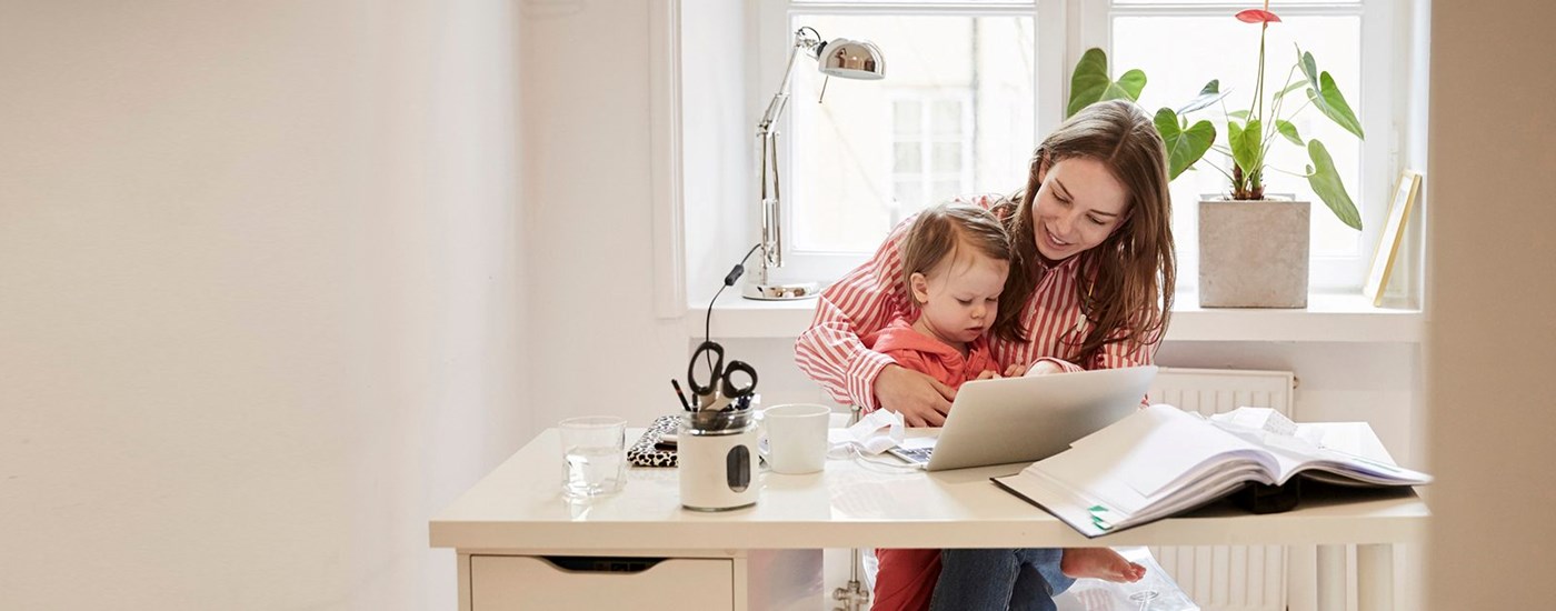 Dame sitter med et barn på fanget mens hun jobber på PC, til illustrasjon for personlig økonomi