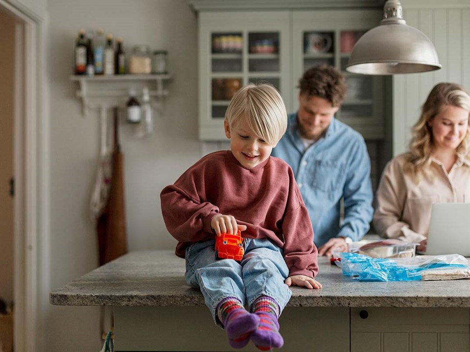 Gutt sitter på kjøkkenbenken og leker med bil med to voksne bak seg