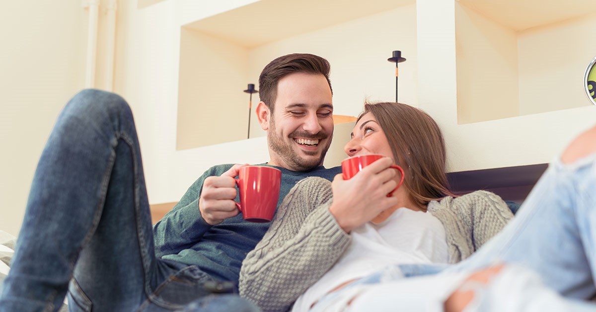 Mann med kaffekopp og kvinne som lener seg på mannen med kaffekopp smiler i sofa, til illustrasjon for slik varer pengene lenger