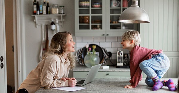 Dame lener seg på kjøkkenbenk med PC ved siden av og et barn som kryper på kjøkkenbenk, til illustrasjon for slik sparer du penger i hverdagen