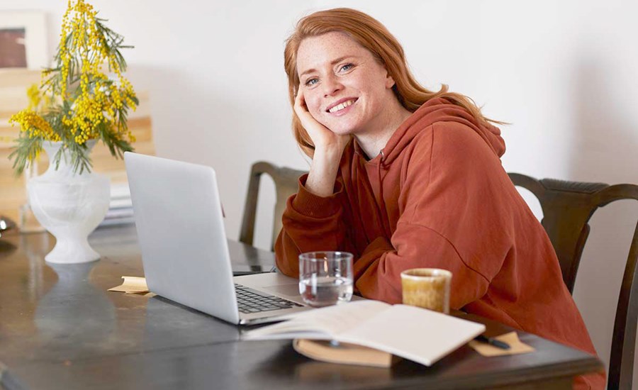 Ung jente sitter på kjøkkenbord med datamaskin fremfor seg og smiler, til illustrasjon for sparekalkulator