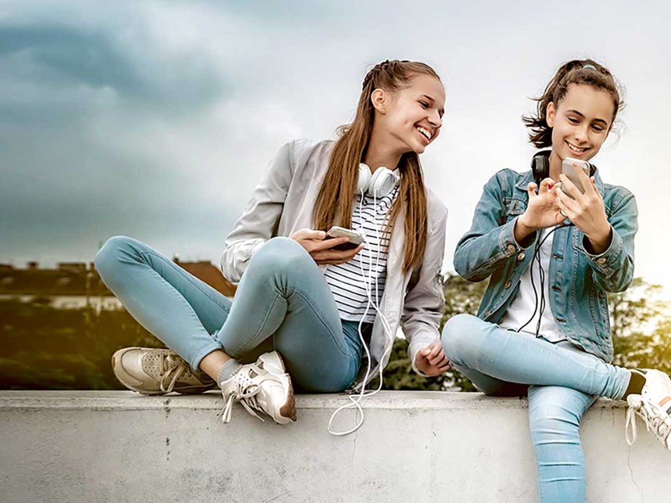To tenåringstjenster med headset rundt halsen og mobilen i handa, til illustrasjon for sparekonto barn og ungdom