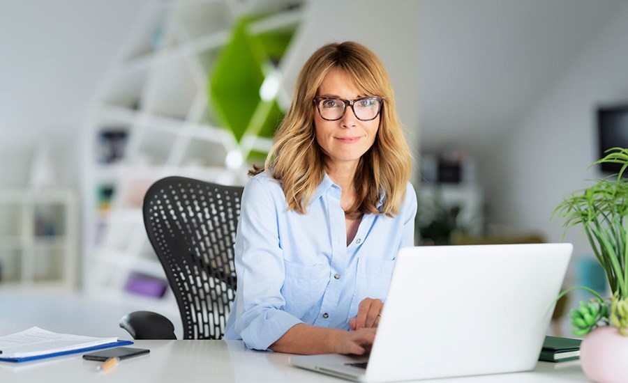 Kvinne med briller og blå skjorte sitter ved en kontorpult og jobber på en laptop, til illustrasjon for betalingstjenester