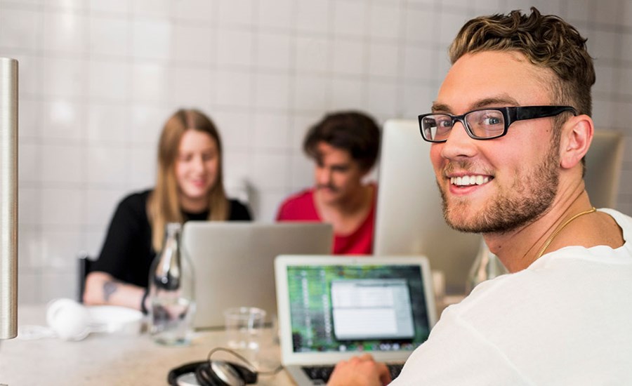Ung mann sitter og jobber på PC mens han smiler med to kollegaer fremfor han, til illustrasjon for kapitalkonto