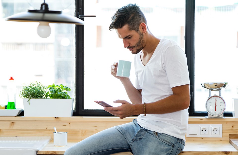 Mann sitter på kjøkkenbenk med kaffe og mobil i hendene