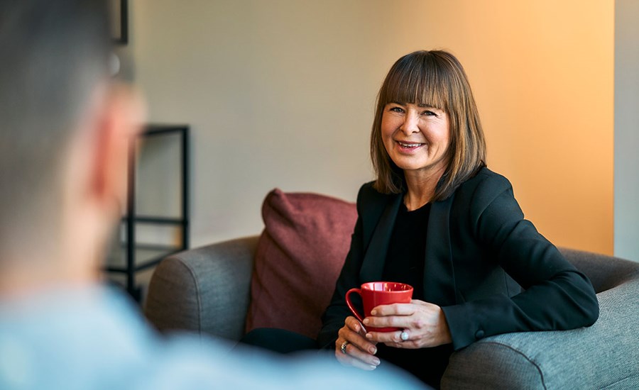 Kvinne sitter i en grå stol og holder en rød kopp mens hun smiler, til illustrasjon for BN Bank