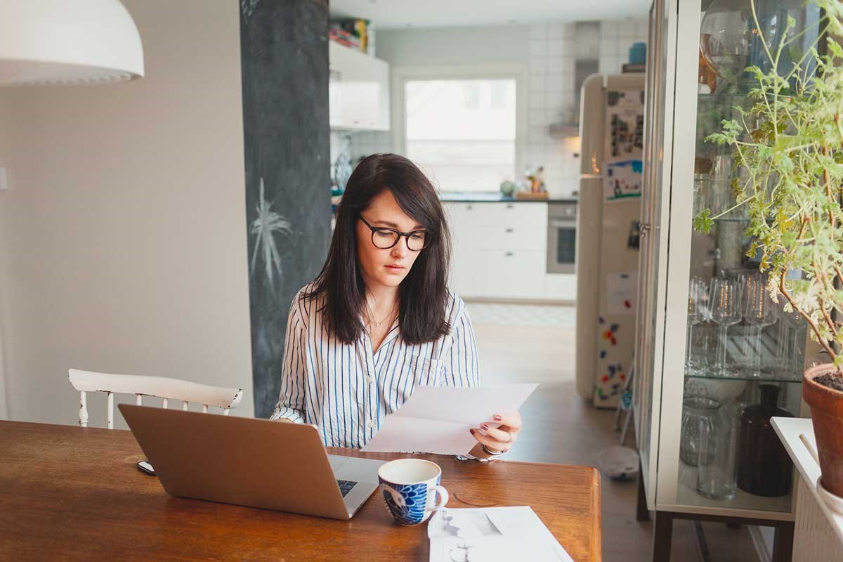Dame sitter på kjøkkenbordet med mobil i hånda og leser dokumenter, til illustrasjon for dødsbo og arveoppgjør
